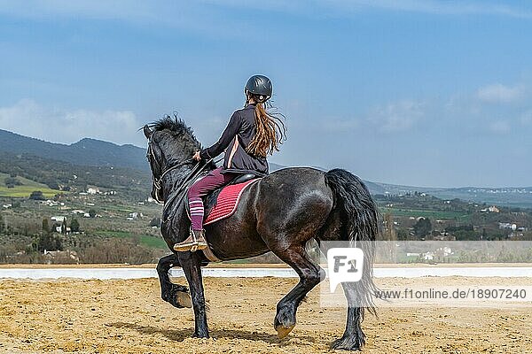 Unerkennbare Frau reitet auf einem schwarzen Pferd mit einer bergigen Landschaft im Hintergrund und blauem Himmel