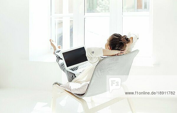 Barfüßige junge Frau mit Händen hinter dem Kopf  die auf einem Stuhl sitzt und sich mit geschlossenen Augen entspannt  nachdem sie einen Laptop in einem hellen Raum zu Hause benutzt hat