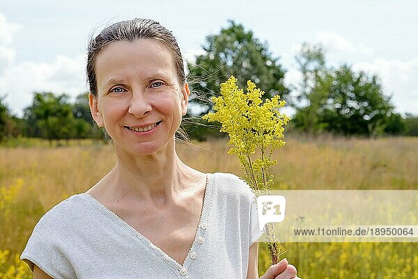 Porträt einer erwachsenen Frau  die in der warmen und weichen Sommersonne auf einer Blumenwiese steht  die auch als Labkraut oder gelbes Labkraut bekannt ist  mit einem Strauß gelber Blüten (Galium verum) in den Händen