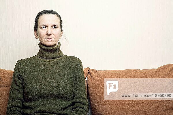 Dramatisches Porträt einer traurigen und depreßiven erwachsenen Frau  die auf einem Sofa sitzt und mit glanzlosen Augen ins Leere schaut. Negativer Raum für Text