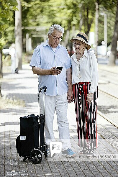 Älteres Paar wartet mit einem Smartphone in der Hand an einer Haltestelle auf die Straßenbahn  Köln  Nordrhein-Westfalen  Deutschland  Europa