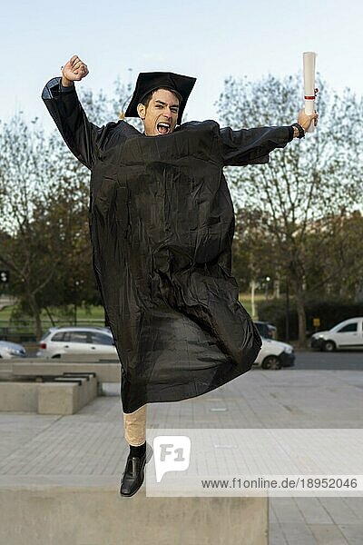Ein Mann  der vor kurzem seinen Abschluss gemacht hat  in Kappe und Talar  zeigt springend seinen Abschluss und feiert