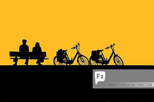 Ein Paar älterer Radfahrer  die sich auf einer Bank neben ihren beiden Fahrrädern ausruhen  silhouettiert gegen den gelben Sonnenuntergangshimmel im Sommer