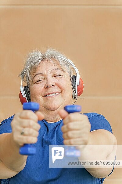 Frontalansicht einer fettleibigen weißhaarigen älteren Frau  die mit Hanteln und Kopfhörern trainiert und Musik hört. lächelnd