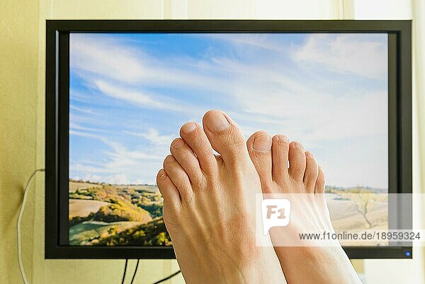 Die Füße eines Mannes vor einem Computermonitor mit einer schönen sonnigen italienischen Landschaft mit bewölktem Himmel  um den Wunsch nach Erholung im Urlaub zu symbolisieren