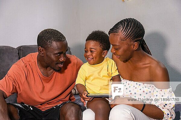 Afrikanische Familie schwarzer Ethnizität mit ihrem Sohn auf dem Sofa zu Hause lächelnd und Spaß mit dem Telefon haben