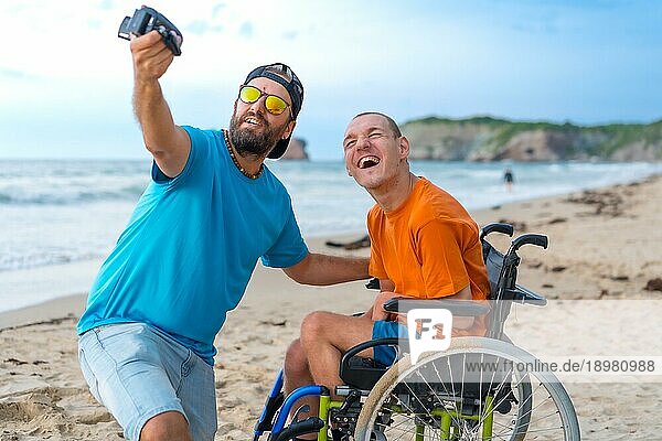 Eine behinderte Person in einem Rollstuhl am Strand mit einem Freund  der ein Selfie macht