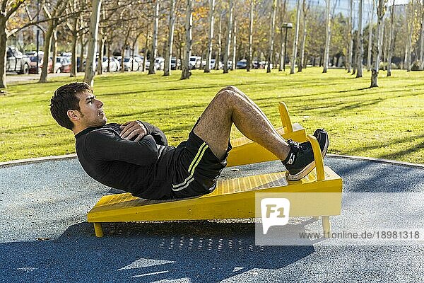 Attraktiver junger Mann bei Bauchmuskelübungen an einem öffentlichen Gerät im Outdoor Fitnessbereich