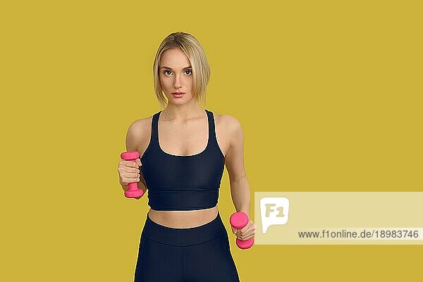 Hübsche blonde Frau in schwarzem Trainingsoberteil mit kleinen rosa Hanteln in den Händen  die in die Kamera schauen. Nahaufnahme Frontalporträt vor einfachem gelben Hintergrund