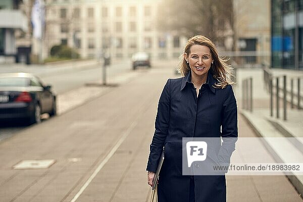 Attraktive  elegante  blonde Frau verlässt ihren Arbeitsplatz und geht lächelnd eine ruhige städtische Straße hinunter  während sie ihren Laptop mit Kopierfunktion mit sich führt