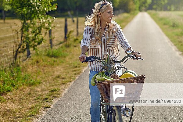 Glückliche Frau lächelt  während sie mit einem Korb voller frischer und gesunder Früchte an einem sonnigen Sommertag auf dem Lande Fahrrad fährt