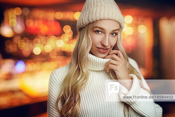 Schöne junge blonde Frau in weißem Strickpulli und Wintermütze  schaut in die Kamera und lächelt  steht vor warmen gelben Lichtern mit Bokeheffekt verschwommen
