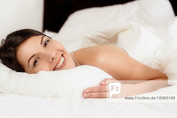 Lächelnde  gesunde  verjüngte junge Frau  die sich im Bett zwischen ihre Kissen kuschelt und der Kamera ein breites  freudiges Lächeln schenkt