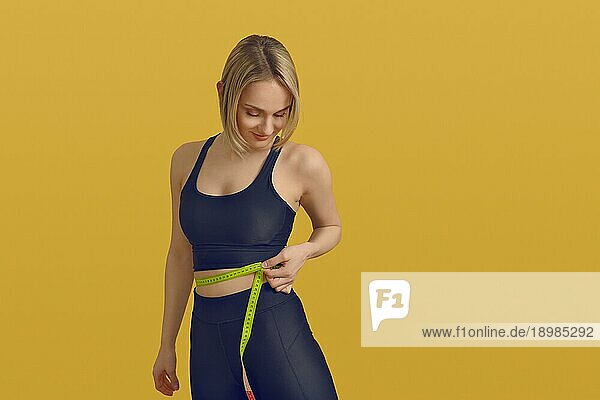 Schlanke  sportliche junge Frau  die ihre Taille mit einem Maßband mißt  während sie mit einem zufriedenen Lächeln nach unten blickt  in einem Konzept für Gewichtsabnahme  gesunde Ernährung und Bewegung auf einem gelben Hintergrund mit Kopierraum