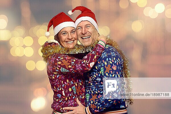 Fröhliches Ehepaar  das Weihnachten zusammen feiert  mit festlichen roten Weihnachtsmannmützen und Weihnachtsmützen  die sich umarmen und lachen  während sie sich umdrehen und in die Kamera lächeln  vor einem Bokeh aus Partylichtern