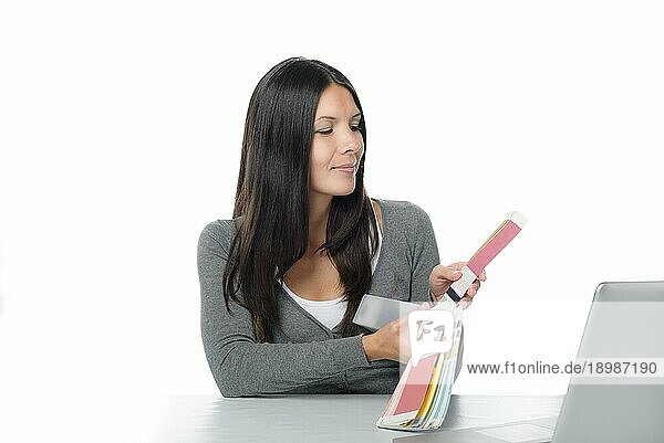 Junge Frau vergleicht online auf ihrem Laptop Farben mit einem Farbmuster  das sie mit einem zufriedenen Lächeln in der Hand hält  auf Weiß