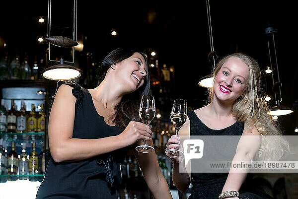 Zwei schöne junge Frauen in eleganten schwarzen Kleidern  die mit alkoholischen Getränken in einer schicken Location feiern