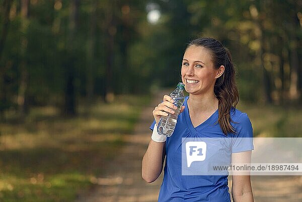 Fitte schlanke junge Frau in Sportkleidung trinkt Wasser in Flaschen  während sie auf einem Waldweg eine Pause einlegt  um während eines Trainingslaufs auf dem Lande neue Flüssigkeit zu sich zu nehmen  Gesundheits und Fitnesskonzept