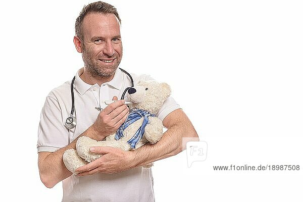 Lächelnde  unrasierte  fürsorgliche Kinderkrankenschwester oder Kinderarzt mit einem Teddybären im Arm  der seine Temperatur mit einem Thermometer mißt  um einen kleinen Patienten während einer Konsultation zu beruhigen