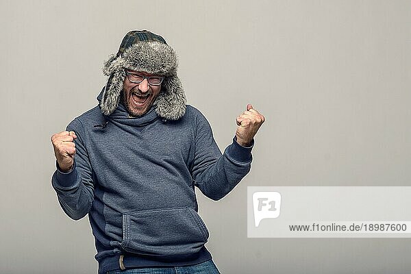 Fröhlicher Mann mit Brille und Wintermütze  der jubelnd und feiernd die geballten Fäuste in die Luft streckt  mit jubelndem Gesichtsausdruck  über grauem Hintergrund mit Kopierraum