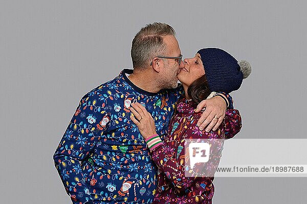 Glückliches Paar mittleren Alters  das sich küsst  während es zusammen Weihnachten feiert und in bunter  weihnachtlicher Kleidung vor einer grauen Wand posiert