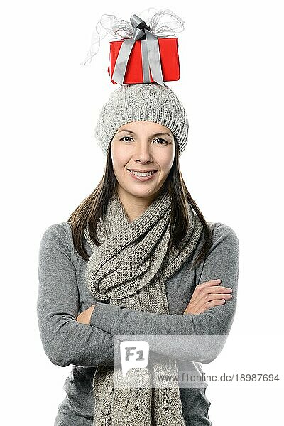 Glückliche  attraktive junge Frau in Wintermode  die ein Weihnachtsgeschenk auf dem Kopf balanciert und in die Kamera schaut  die Hände vor sich verschränkt und mit einem charmanten Lächeln  vor weißem Hintergrund