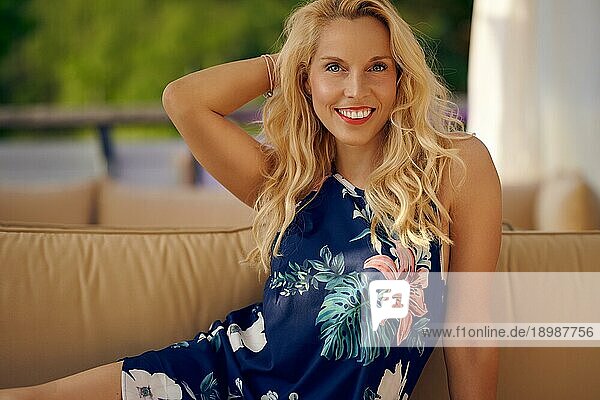 Lächelnde  attraktive  freundliche  blonde Frau  die sich in einem bunten  geblümten Sommerkleid auf einem Sofa ausruht  in einer Nahaufnahme und glücklich in die Kamera grinsend