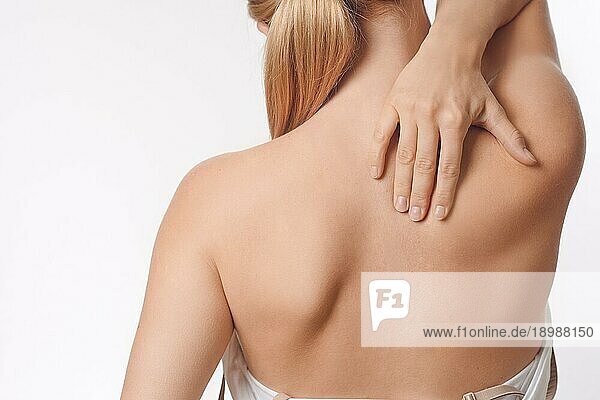 Frau mit Schmerzen im oberen Rücken und Nacken  die nackt mit dem Rücken zur Kamera steht und mit der Hand ihre Schultermuskeln nahe der Wirbelsäule reibt