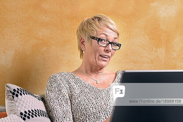 Ernste Frau mittleren Alters mit kurzen blonden Haaren  die auf dem Sofa sitzt und ihren Laptop benutzt