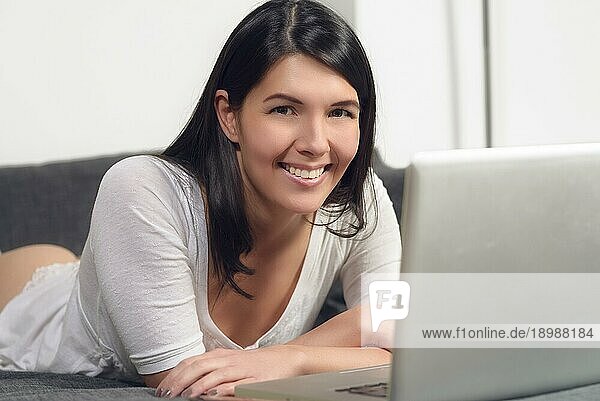Attraktive junge Frau  die mit ihrem Laptop auf dem Sofa liegt und eine Pause einlegt und der Kamera ein charmantes  freundliches Lächeln schenkt