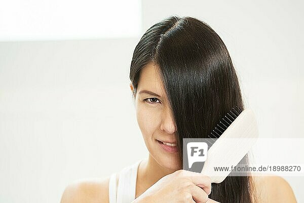 Attraktive Frau  die sich die Haare bürstet  wobei ein Auge von ihren langen brünetten Locken verdeckt wird und sie mit einem sinnlich verführerischen Blick in die Kamera schaut
