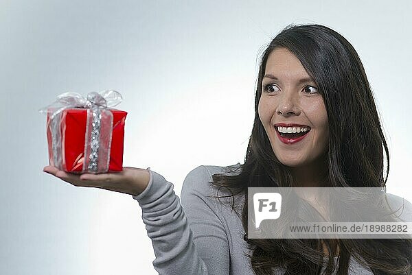 Schöne junge Frau  die ein dekoratives rotes Geschenk betrachtet  das sie in Aufregung und Vorfreude auf die Überraschung in ihrer Handfläche hält