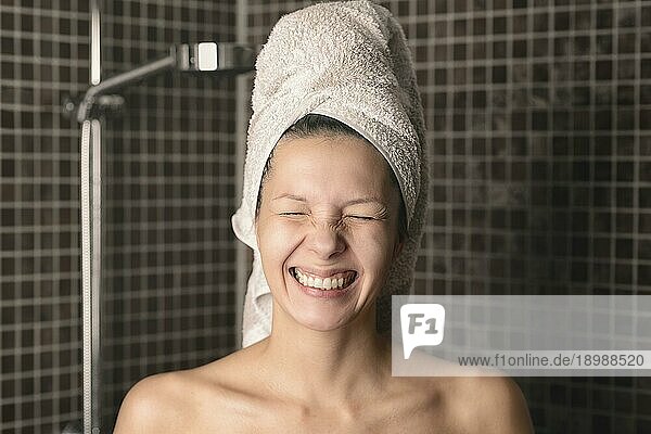Verspielte  verschmitzte  nackte Frau mit naßen Haaren in einem Handtuch  die mit geschlossenen Augen in die Kamera grinst  während sie im Badezimmer steht  offenes Porträt
