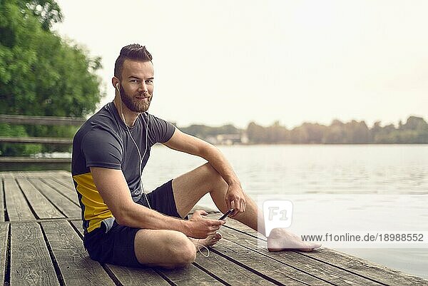 Attraktiver barfüßiger  bärtiger junger Mann  der sich auf einer Holzterrasse mit Blick auf einen ruhigen See entspannt  sein Handy in der Hand und mit einem Lächeln in die Kamera schaut