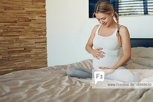 Glückliche junge schwangere Frau  die mit ihrem ungeborenen Kind auf einem Bett sitzt und ihren Babybauch mit ihren Händen wiegt und mit einem zärtlichen Lächeln der Liebe nach unten schaut