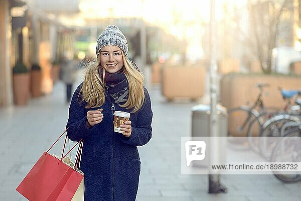 Wunderschöne junge Frau mit Weihnachtseinkäufen in einer gestrickten Wintermütze  die fröhlich lächelt  während sie auf einer städtischen Straße hinter sich blickt