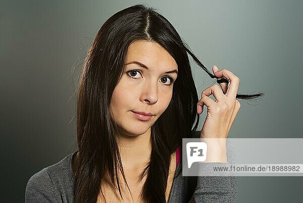 Nachdenkliche attraktive junge Frau  die ihr langes brünettes Haar um ihre Finger zwirbelt  während sie mit einem wachsamen  nachdenklichen Ausdruck in die Kamera schaut