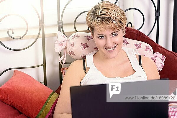 Junge blonde Frau mit Laptop  die sich auf Kissen auf dem Bett stützt und in die Kamera schaut  von oben