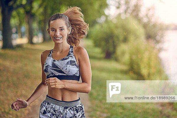 Fitte  gesunde  sportliche Frau  die an einem Flussufer joggt und dabei lächelnd ihr langes Haar nach hinten wirft  während sie sich der Kamera nähert