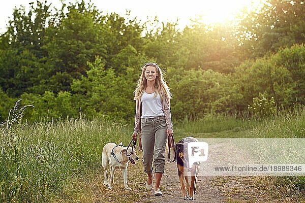 Glückliche junge Frau  die mit ihren Hunden auf einem grasbewachsenen Feldweg im Frühling spazieren geht und in die Kamera schaut