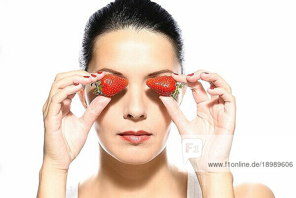 Schöne junge Frau mit einem schönen warmen Lächeln und gesunder gebräunter Haut  die zwei reife rote Erdbeeren an die Seiten ihrer Augen hält  vor weißem Hintergrund