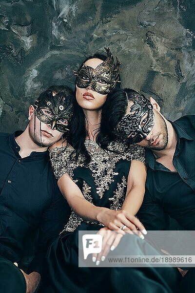 Schöne junge Frau und zwei Männer in mysteriösen schwarzen Masken sitzen auf dem Boden in einem Studio. Mode  Party Konzept