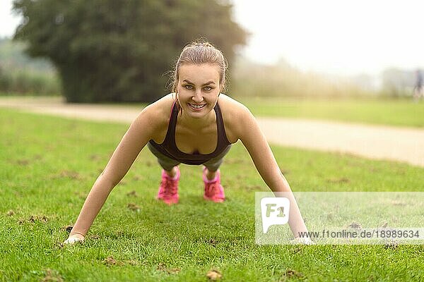 Sportliche junge Frau lächelt in die Kamera  während sie im Park Liegestützübungen macht