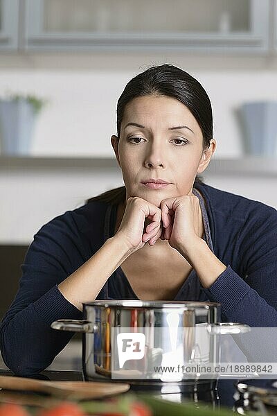Unmotivierte attraktive junge Frau  die das Abendessen zubereitet und sich dabei auf das Kochfeld stützt und das frische Gemüse mit lustlosem  mürrischem Blick betrachtet  während sie in ihrer Küche in einer Schürze steht