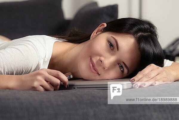 Attraktive junge Frau  die ihren Kopf auf einen Laptop stützt  während sie sich auf dem Sofa ausruht und der Kamera ein charmantes  freundliches Lächeln schenkt