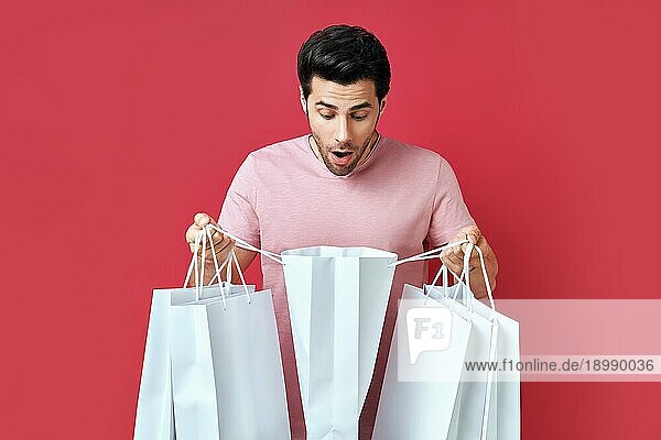 Junger verwirrter Mann  der Einkaufstaschen in den Händen hält und seinen Einkauf vorrotem Hintergrund betrachtet. Einkaufen  Verkauf  shopaholic Konzept