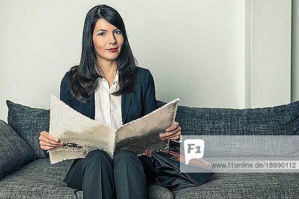 Attraktive Geschäftsfrau im eleganten Schlabberanzug  die auf einem grauen Sofa sitzt und eine Geschäftszeitung oder eine Zeitung liest  während sie mit ihrer Aktentasche neben sich auf einen Termin wartet