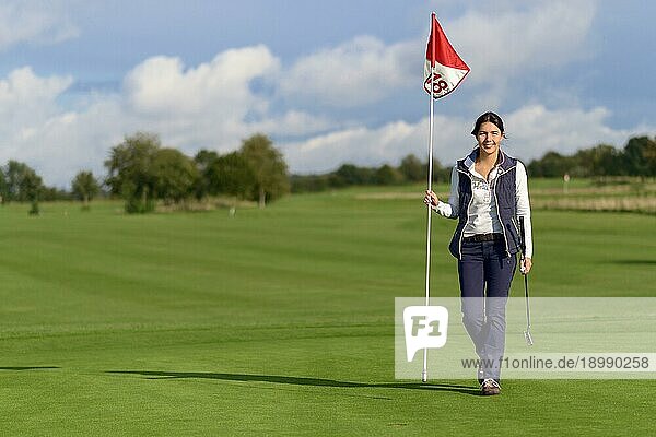 Eine Golferin hält die Fahne des Lochs in der Hand  als sie sie entfernt  um ihren letzten Putt auf dem Grün zu machen  und lächelt in die Kamera