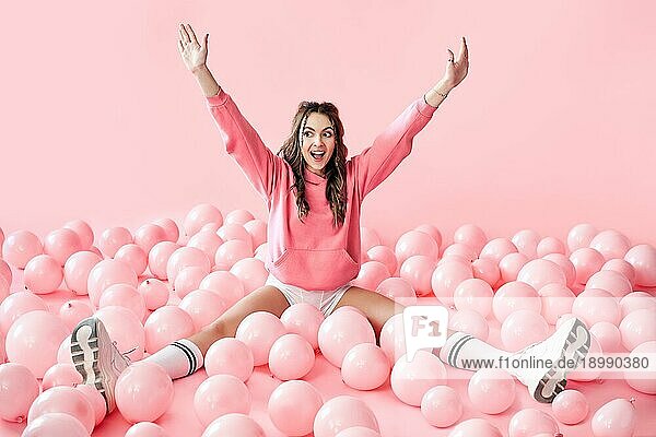 Junge glückliche Frau mit den Händen nach oben sitzen in rosa Luftballons auf Pastell rosa Hintergrund. gewinnen  Feier  Spaß Konzept