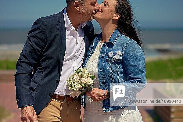 Glückliches  romantisches  reifes  frisch verheiratetes Paar an ihrem Hochzeitstag  das sich im Freien auf einer Promenade vor einer Meereskulisse küsst  formelle Kleidung trägt und einen Blumenstrauß hält